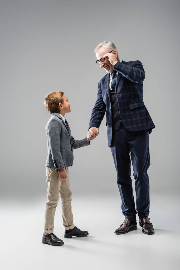 homem de negócios feliz tocando óculos enquanto segurando as mãos com o neto no cinza - Foto, Imagem