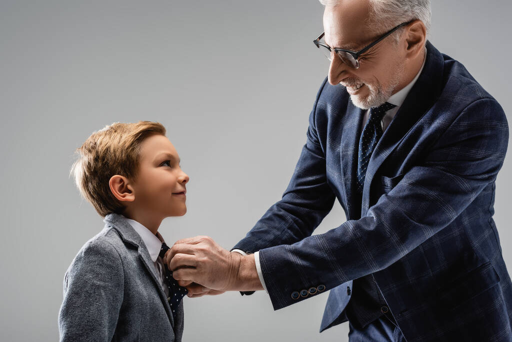 gelukkig zakenman in bril knopen blazer van glimlachende kleinzoon geïsoleerd op grijs - Foto, afbeelding
