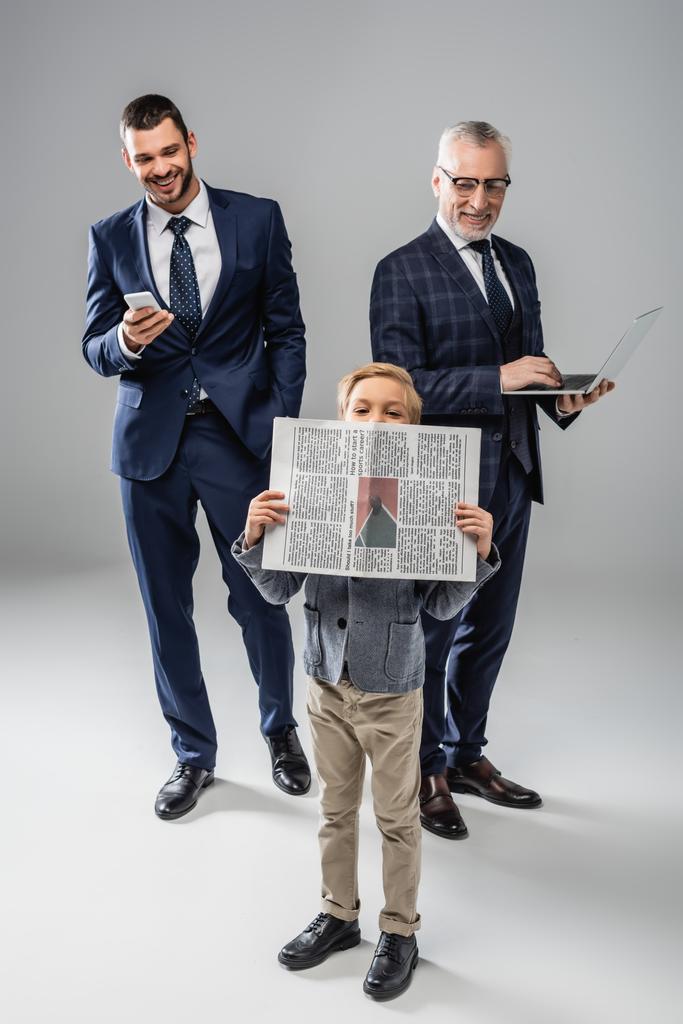 sonrientes hombres de negocios en ropa formal sosteniendo gadgets y mirando a chico con periódico en gris - Foto, Imagen
