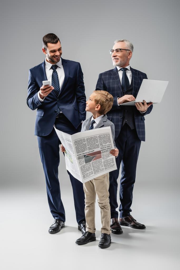 garçon souriant avec journal près de papa et grand-père tenant gadgets sur gris - Photo, image
