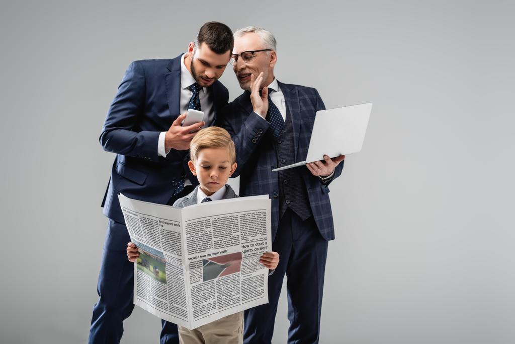 зрелый бизнесмен разговаривает с сыном рядом мальчик в формальной одежде читает газету изолированы на серый - Фото, изображение