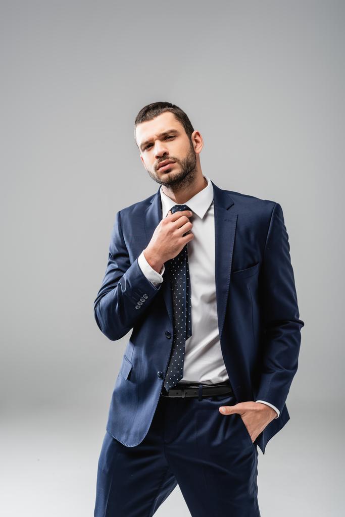 selbstbewusster Geschäftsmann fixiert Krawatte im Stehen mit der Hand in der Tasche auf grau - Foto, Bild