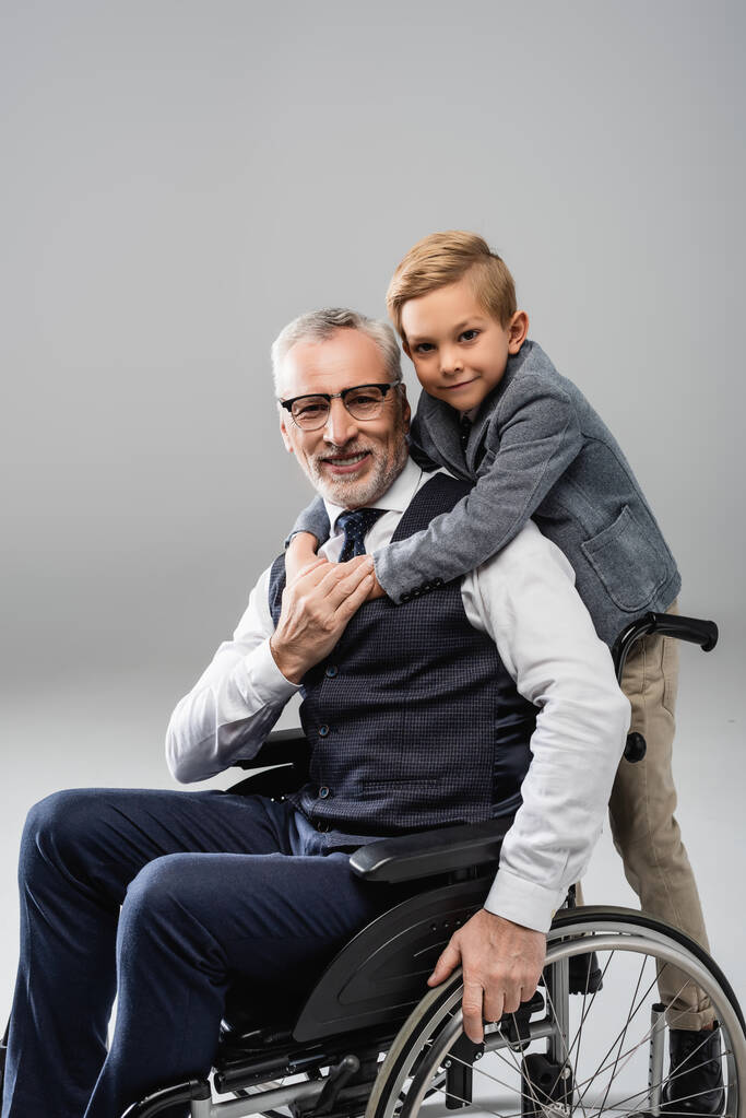 niño sonriente abrazando al abuelo en silla de ruedas mientras miran a la cámara juntos en gris - Foto, imagen