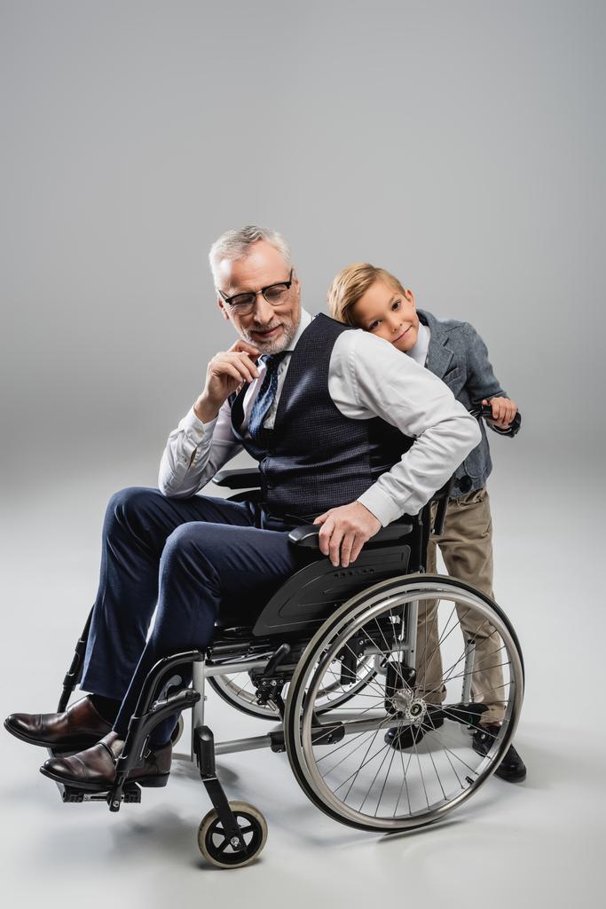 Улыбающийся мальчик опирается на спину дедушки в инвалидном кресле на сером - Фото, изображение