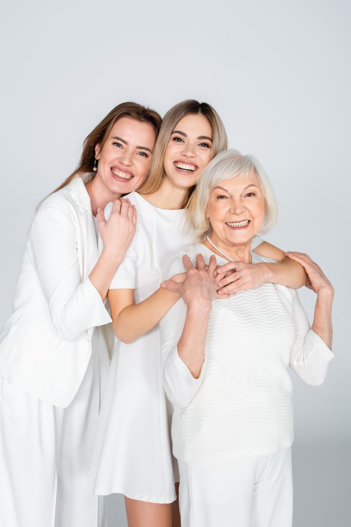 tres generaciones de mujeres sonrientes mirando a la cámara y abrazándose aisladas en gris - Foto, Imagen
