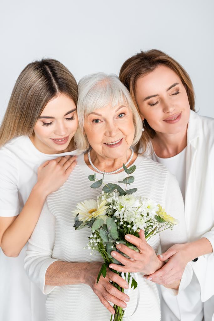 щаслива старша жінка посміхається, тримаючи квіти біля дочки та онуки ізольовані на білому
 - Фото, зображення