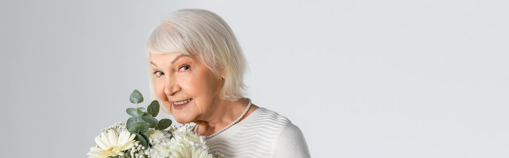 gelukkig senior vrouw met grijs haar houden bloemen geïsoleerd op grijs, banner - Foto, afbeelding