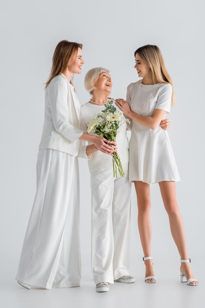 full length of happy senior woman holding flowers and smiling with daughter and granddaughter on grey - Photo, Image