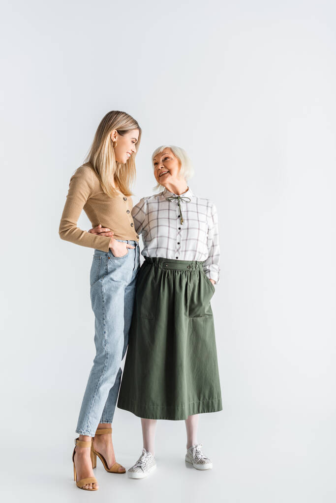 volle Länge der fröhlichen Enkelin und Senior Oma stehend mit den Händen in den Taschen auf weiß - Foto, Bild