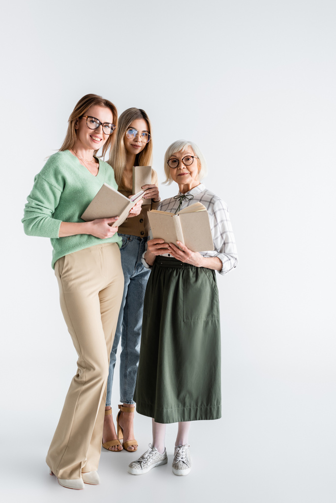 longitud completa de tres generaciones de mujeres sonrientes en gafas con libros en blanco - Foto, Imagen