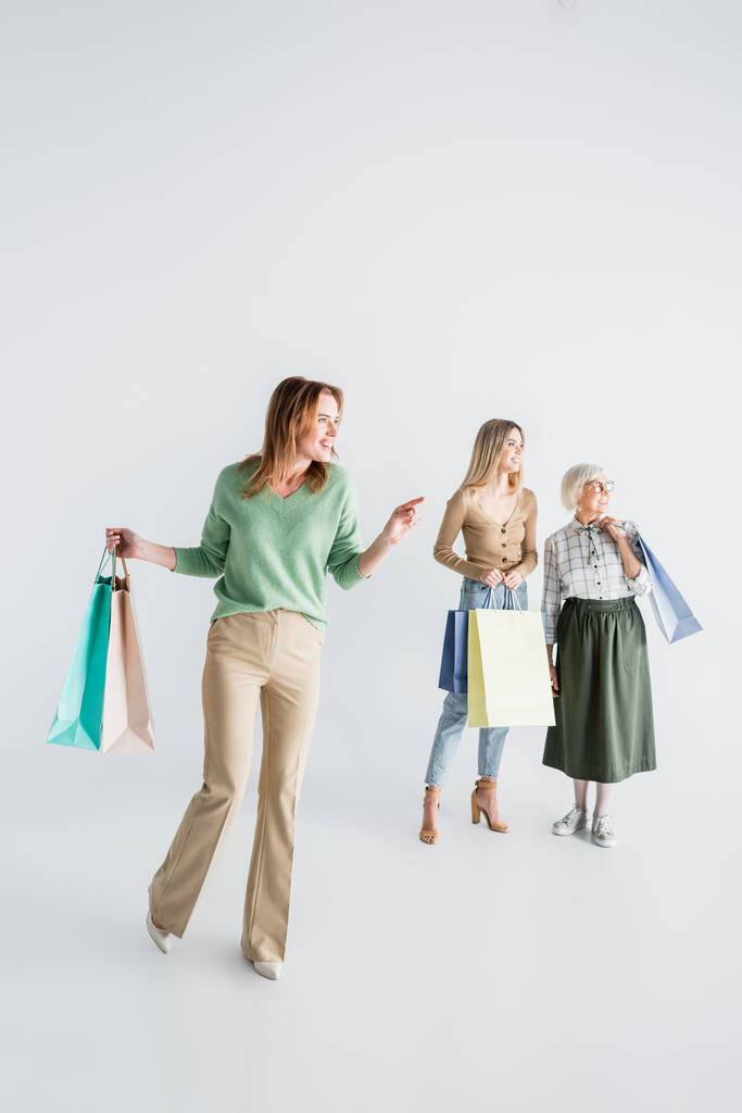 Elinde alışveriş poşetleri olan mutlu bir kadın ve bulanık beyaz arka planda kızı ve yaşlı annesini işaret ediyor. - Fotoğraf, Görsel