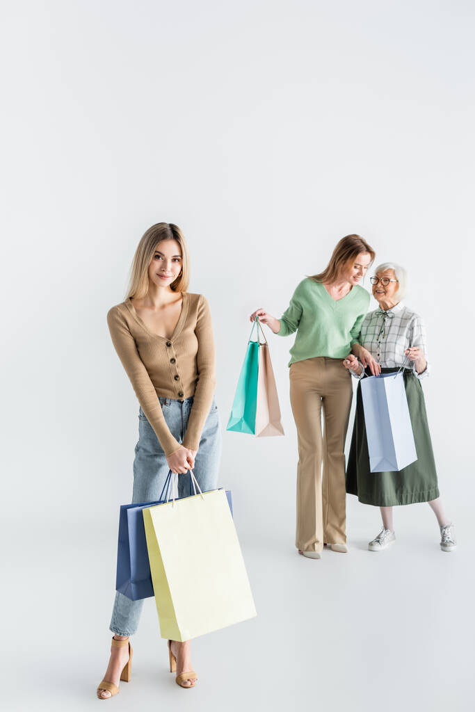 幸せな若い女性の完全な長さで立っていますショッピングバッグとともにお母さんとおばあちゃんの近くにぼやけた背景 - 写真・画像