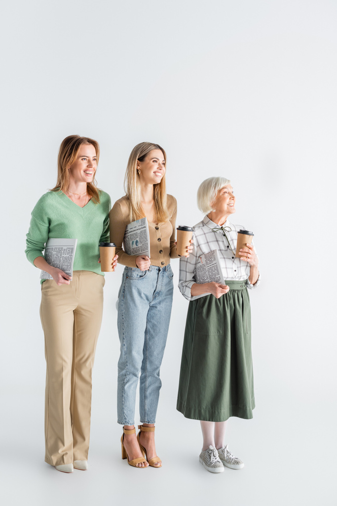 full length of three generation of happy women holding newspapers and paper cups on white - Photo, Image