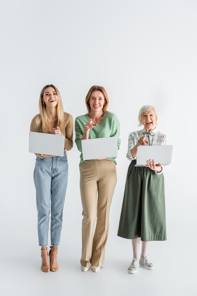 plná délka tří generací veselých žen ukazujících prsty a držících notebooky na bílém - Fotografie, Obrázek
