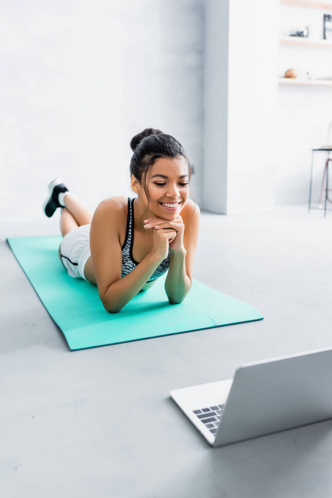 smiling african american sportswoman looking at laptop while lying on fitness mat - Photo, Image