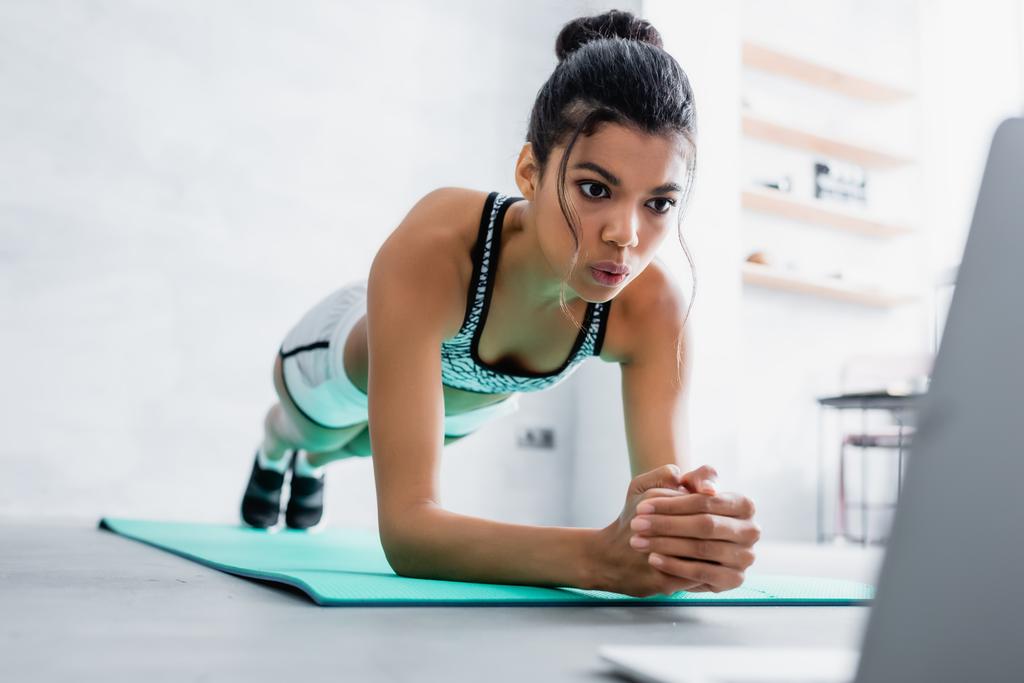 african american sportswoman practicing plank pose while looking at laptop, blurred background - Photo, Image