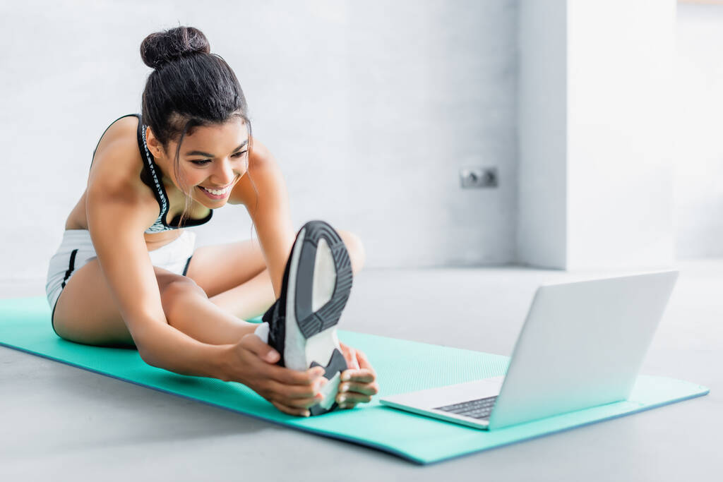 vidám afro-amerikai nő nyújtózkodik, miközben ül a fitness mat közelében laptop - Fotó, kép