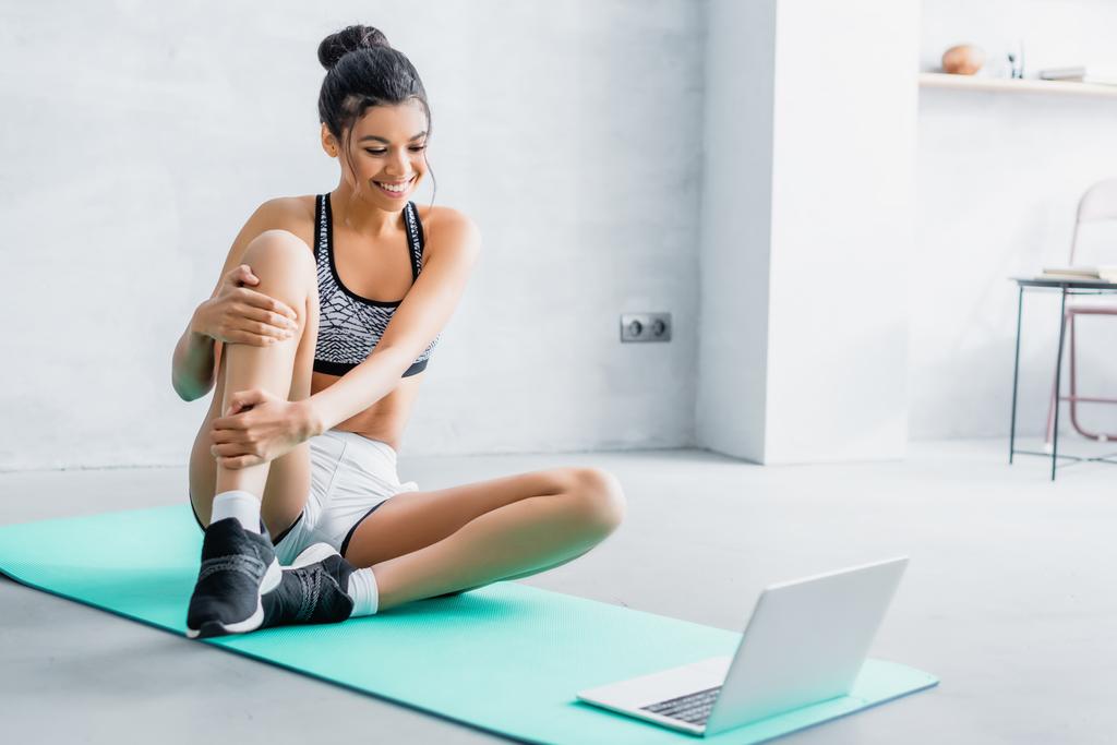 young african american woman in sportswear smiling while sitting on fitness mat near laptop - Photo, Image