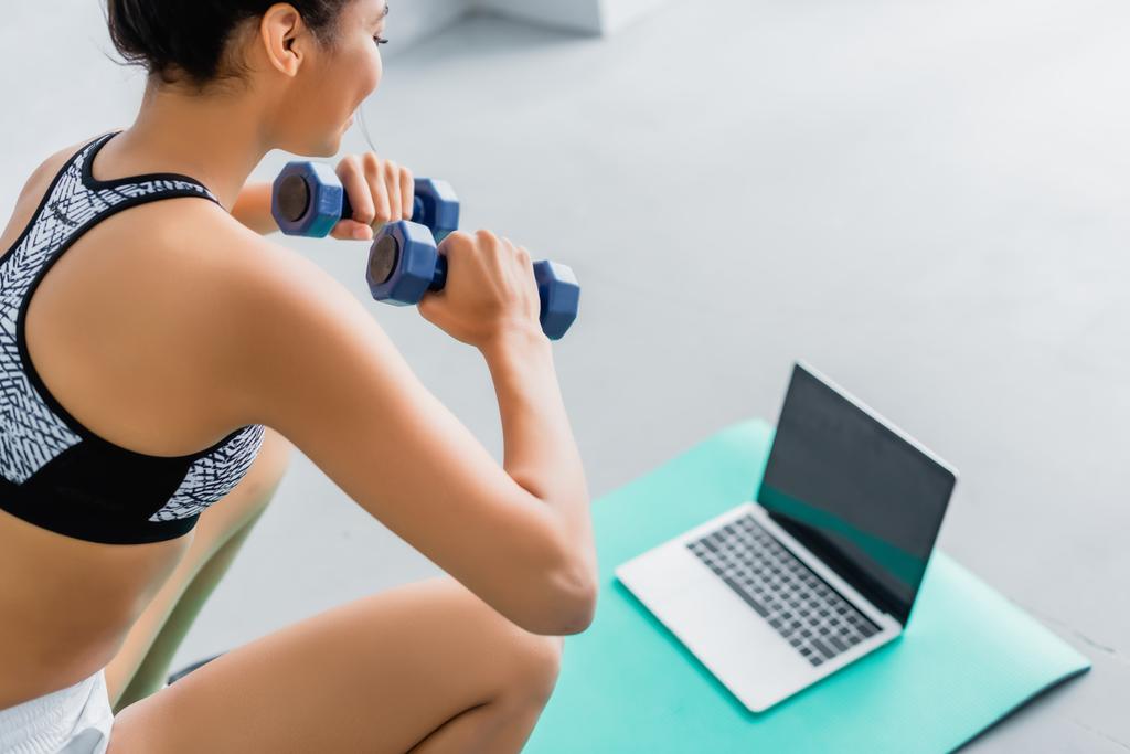 vista recortada de la joven deportista afroamericana haciendo ejercicio con pesas cerca de un ordenador portátil borroso con pantalla en blanco - Foto, Imagen