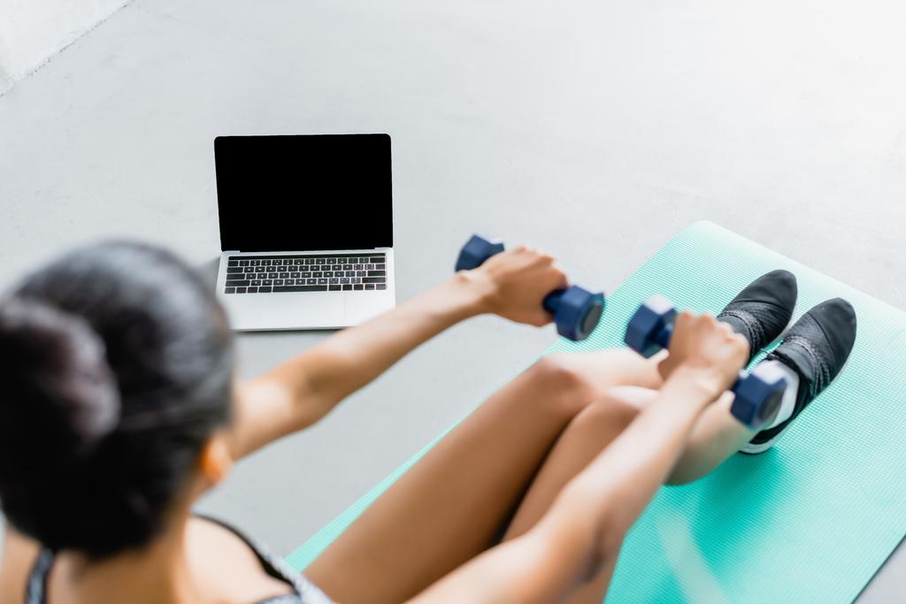 vista aérea de la deportista afroamericana haciendo ejercicio con pesas cerca de la computadora portátil con pantalla en blanco, primer plano borroso - Foto, Imagen