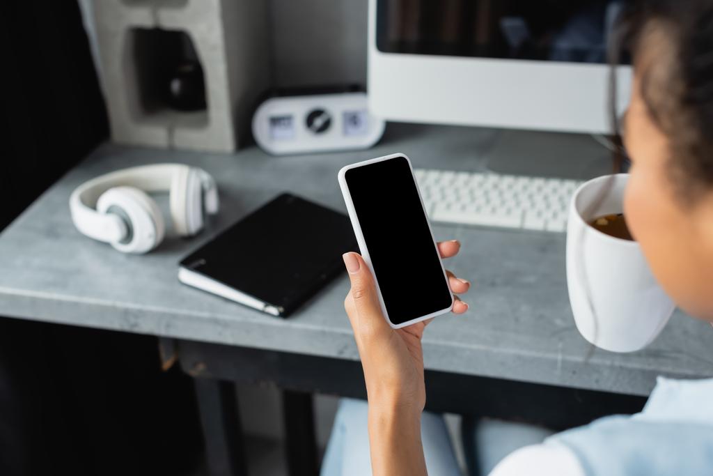 vista ritagliata di afro-americano freelance con smartphone con schermo bianco vicino notebook e cuffie su sfondo sfocato - Foto, immagini