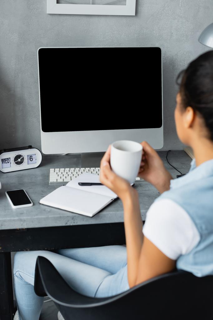 giovane freelance africano americano che tiene una tazza di tè vicino al monitor del computer, smartphone e notebook sulla scrivania, offuscata in primo piano - Foto, immagini