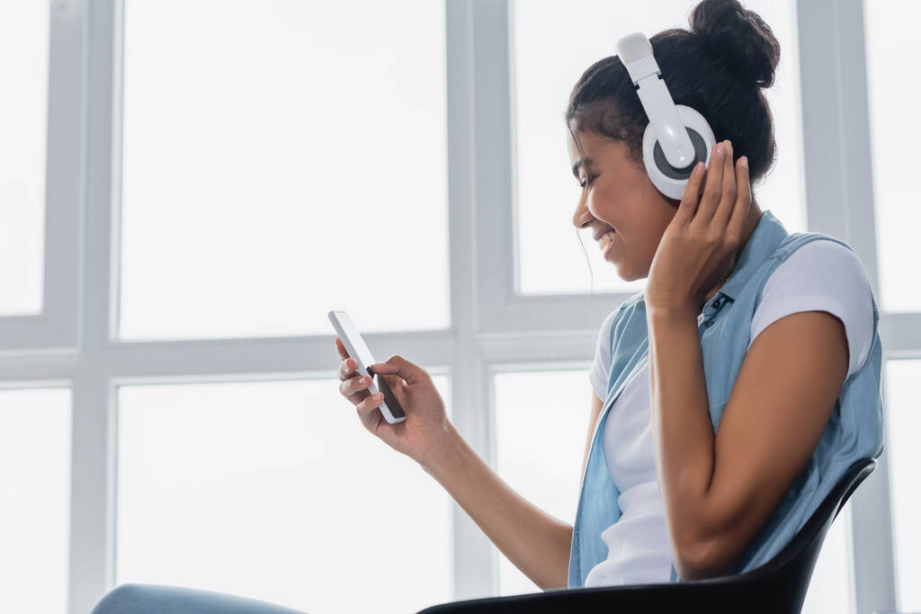 widok z boku Afryki amerykanka czatuje na smartfonie podczas słuchania muzyki w bezprzewodowych słuchawkach w pobliżu okna - Zdjęcie, obraz