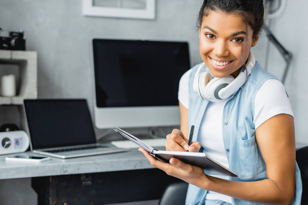 cheerful african american freelancer looking at camera and writing in notebook near laptop and computer monitor on blurred background - Photo, Image