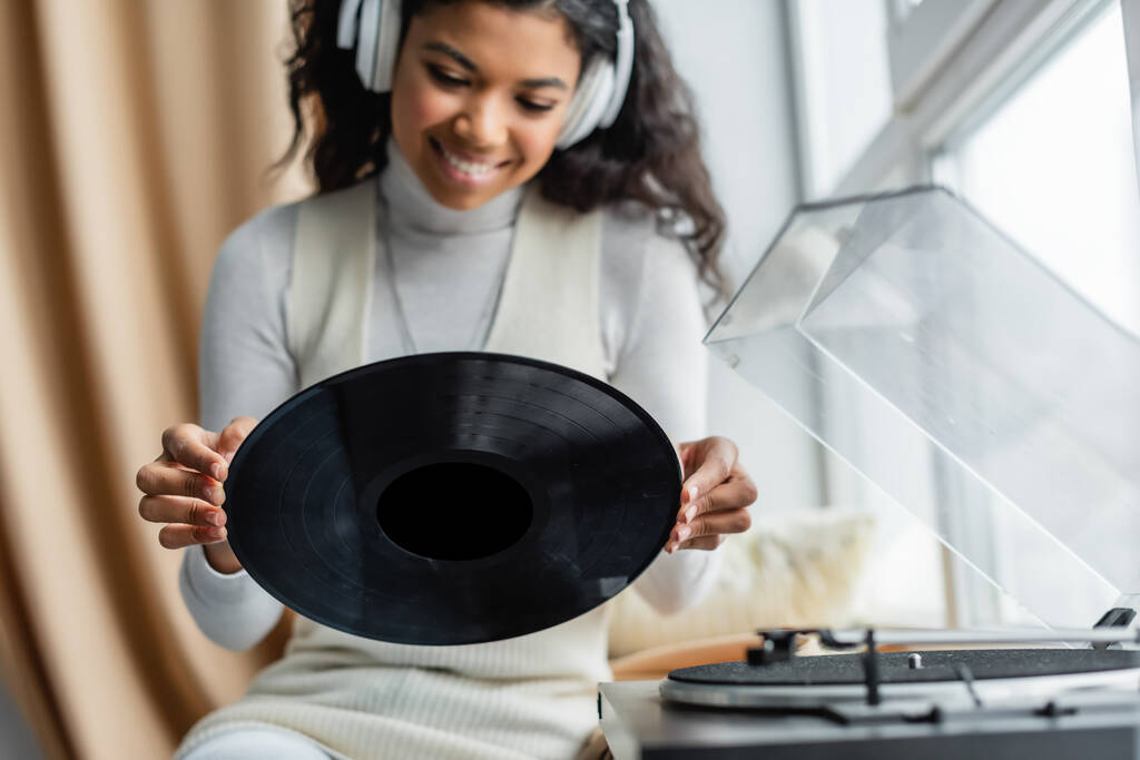 レコードプレーヤーの近くにビニールディスクを保持している無線ヘッドフォンで幸せなアフリカ系アメリカ人女性,ぼやけた前景 - 写真・画像