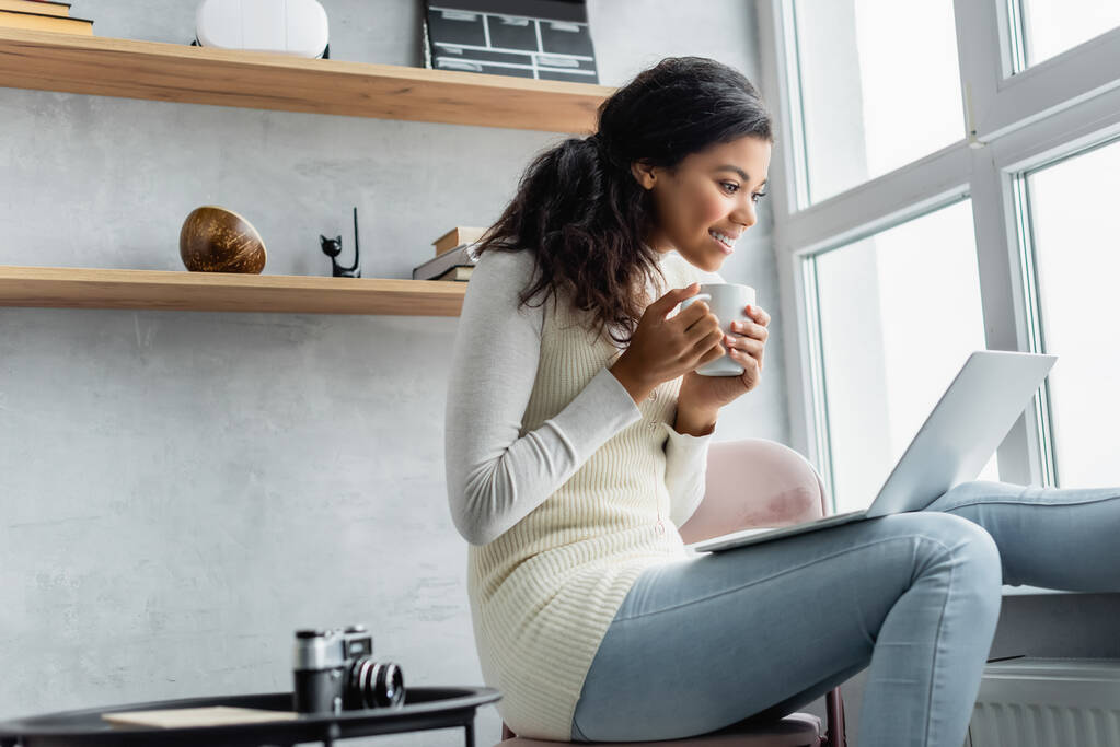 alegre mujer afroamericana con taza de té mirando a la computadora portátil mientras está sentado cerca de la cámara retro en primer plano borrosa - Foto, imagen