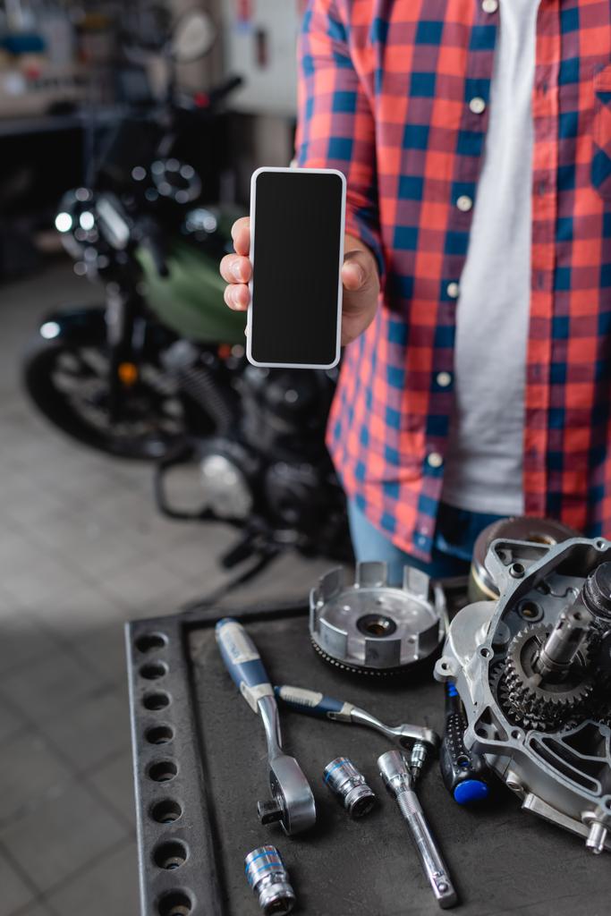 vista parcial del mecánico que muestra el teléfono móvil con la pantalla en blanco cerca de piezas de repuesto y llaves de motocicleta, fondo borroso - Foto, imagen