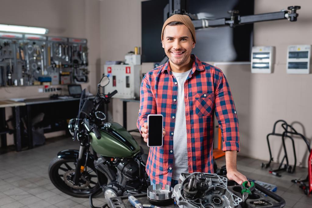веселый механик показывает мобильный телефон с чистым экраном рядом с запасными частями и мотоциклом в мастерской - Фото, изображение
