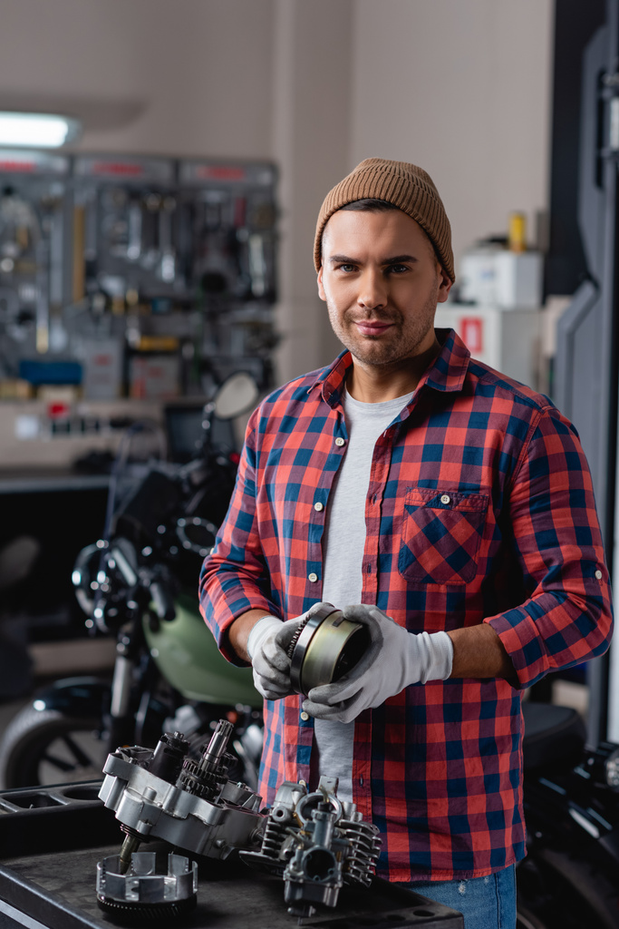молодой механик в клетчатой рубашке и шапке смотрит в камеру рядом с разобранной коробкой передач мотоцикла - Фото, изображение