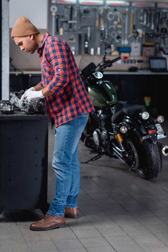 volledige weergave van monteur in geruit shirt en jeans controleren van gedemonteerde versnellingsbak in de buurt van motorfiets in werkplaats - Foto, afbeelding