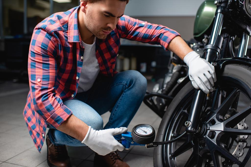 joven mecánico en camisa a cuadros que mide la presión del aire en el neumático de la motocicleta con manómetro - Foto, imagen