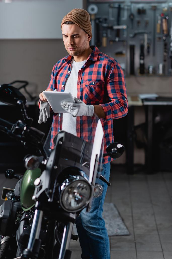 fiatal szerelő sapkában és kockás ingben digitális táblagéppel, miközben a motorkerékpár közelében áll műhelyben - Fotó, kép
