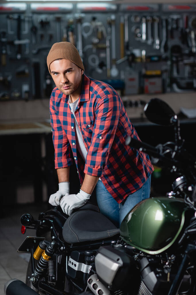 joven reparador en camisa a cuadros y gorro mirando a la cámara mientras está de pie cerca de la motocicleta en el taller - Foto, Imagen