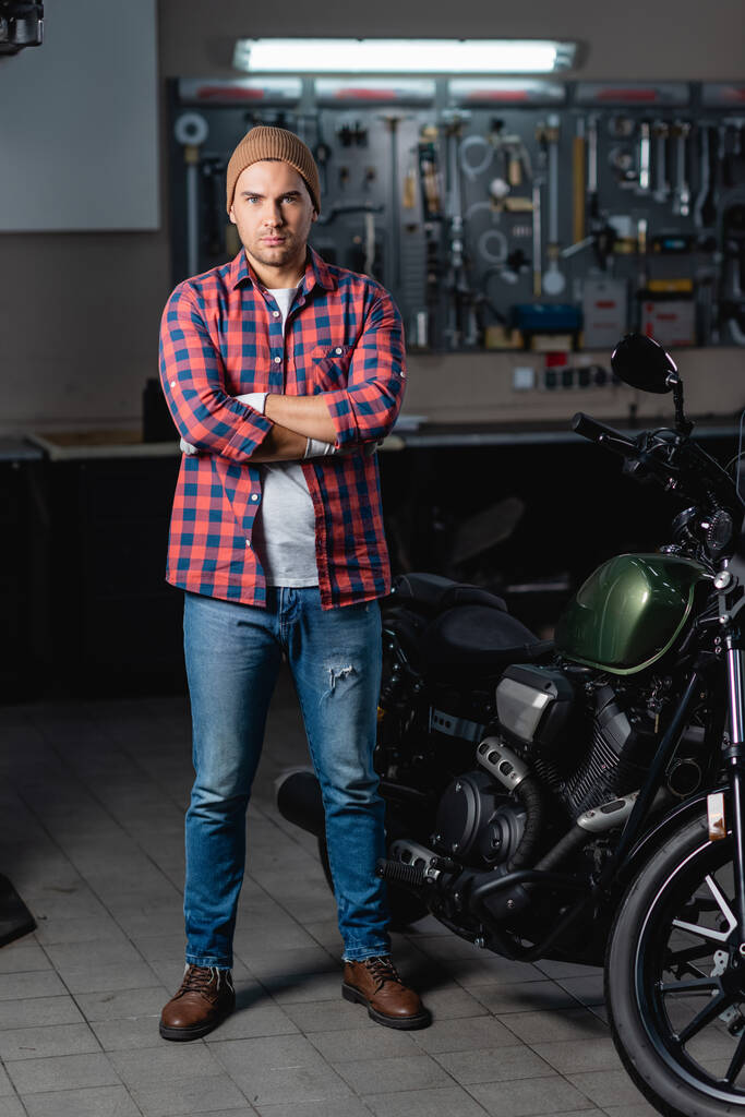volledige weergave van monteur in geruit shirt en jeans met gekruiste armen in de buurt van motorfiets in werkplaats - Foto, afbeelding