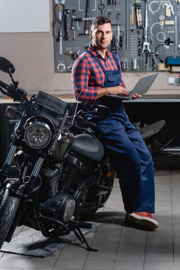 mechanic in overalls looking at camera while standing near motorbike with laptop - Photo, Image