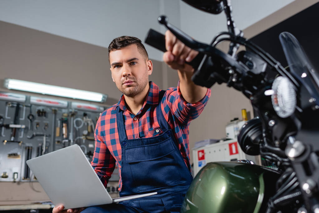 Junger Mechaniker mit Laptop blickt in Werkstatt auf Kamera, während er auf Motorrad sitzt, unscharfer Vordergrund - Foto, Bild