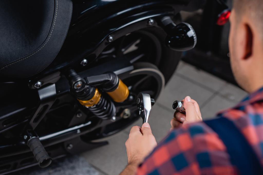 cropped view of mechanic holding socket wrench near motorbike, blurred foreground - Photo, Image