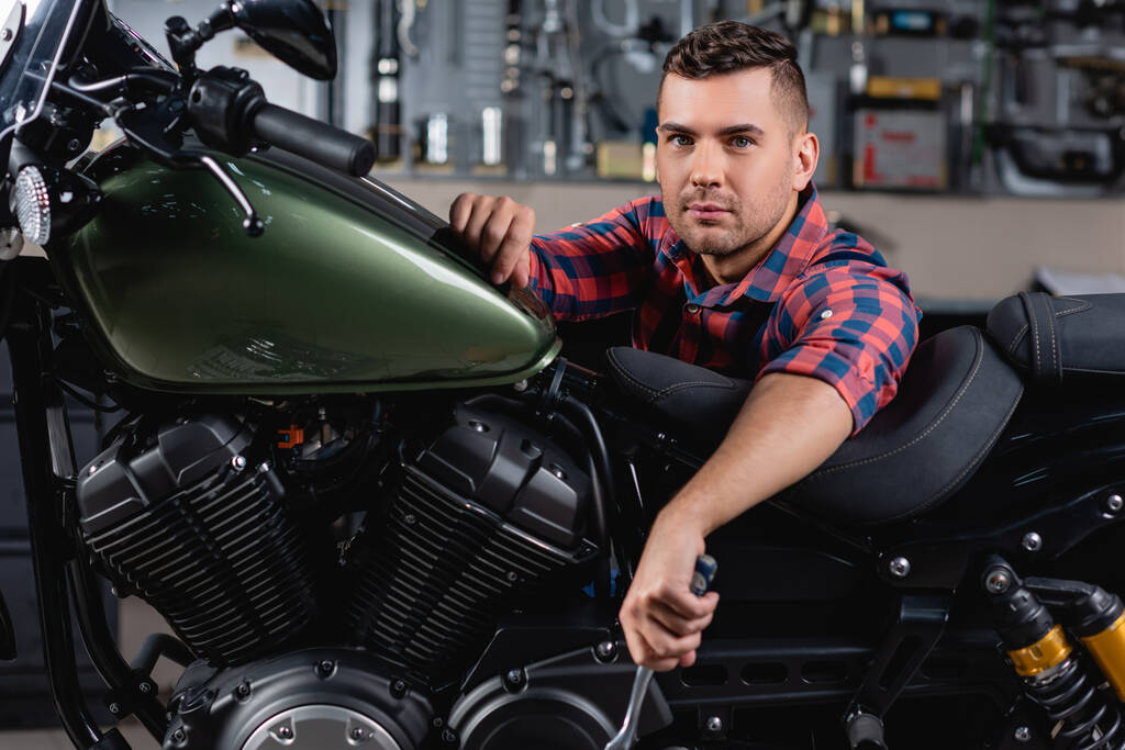 молодой механик смотрит в камеру рядом с мотоциклом в мастерской - Фото, изображение