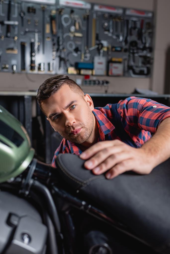 jovem mecânico olhando para a câmera ao fazer diagnósticos de moto em primeiro plano borrado - Foto, Imagem