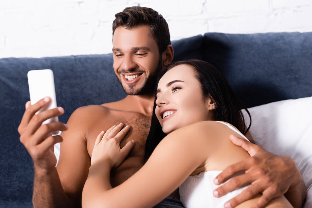 šťastný mladý pár při pohledu na smartphone při ležení na posteli - Fotografie, Obrázek