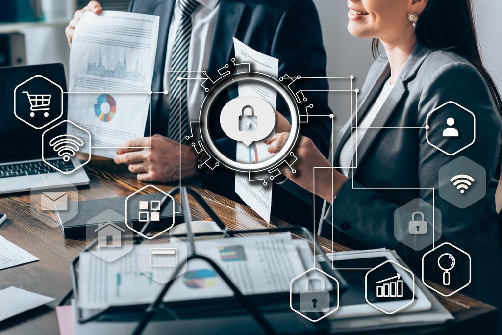 Cropped view of smiling businesswoman holding document with chart near colleague and devices on blurred foreground, lock sign illustration  - Photo, Image