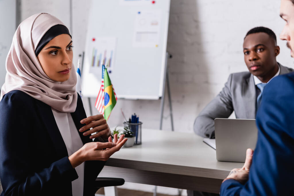 femme d'affaires arabe dans le hijab geste tout en parlant à des partenaires multiculturels lors de la réunion, flou premier plan - Photo, image