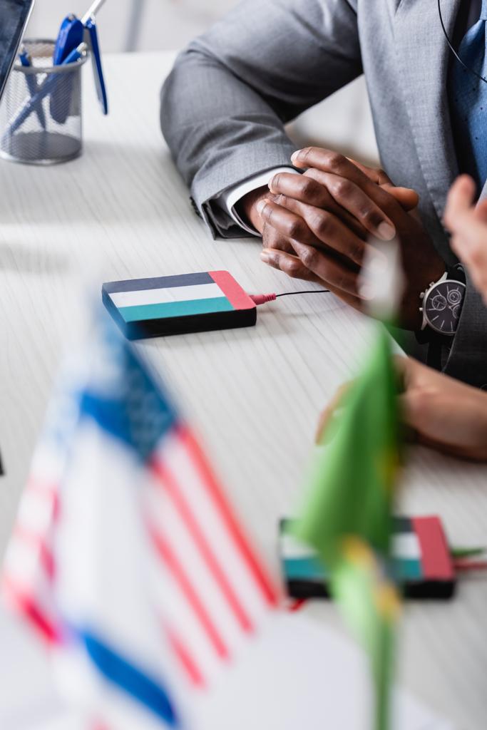 vista cortada do homem de negócios americano africano sentado com as mãos apertadas perto do tradutor digital com o emblema da bandeira dos Estados Unidos, foreground borrado - Foto, Imagem