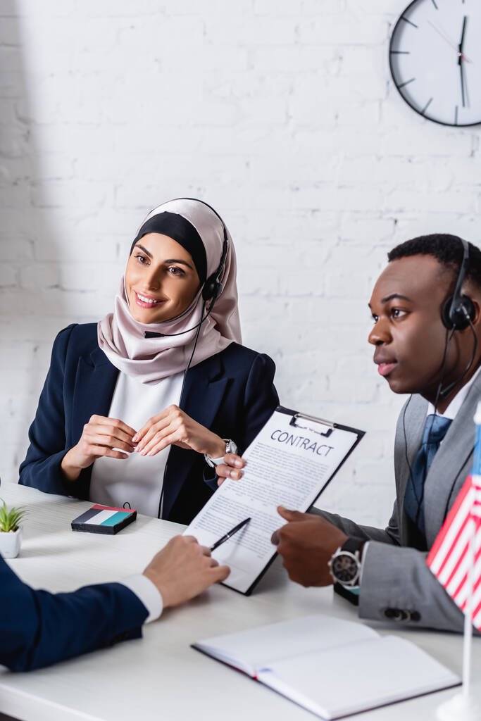 Αραβικοί και αφροαμερικανοί επιχειρηματικοί εταίροι σε ακουστικά κοντά στο μεταφραστή δείχνοντας το συμβόλαιο σε θολά νέα - Φωτογραφία, εικόνα