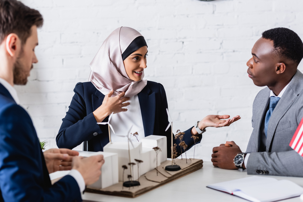sorridente mulher de negócios árabe apontando com a mão enquanto conversa com parceiro de negócios afro-americano perto do modelo de usina de energia verde, foreground borrado - Foto, Imagem