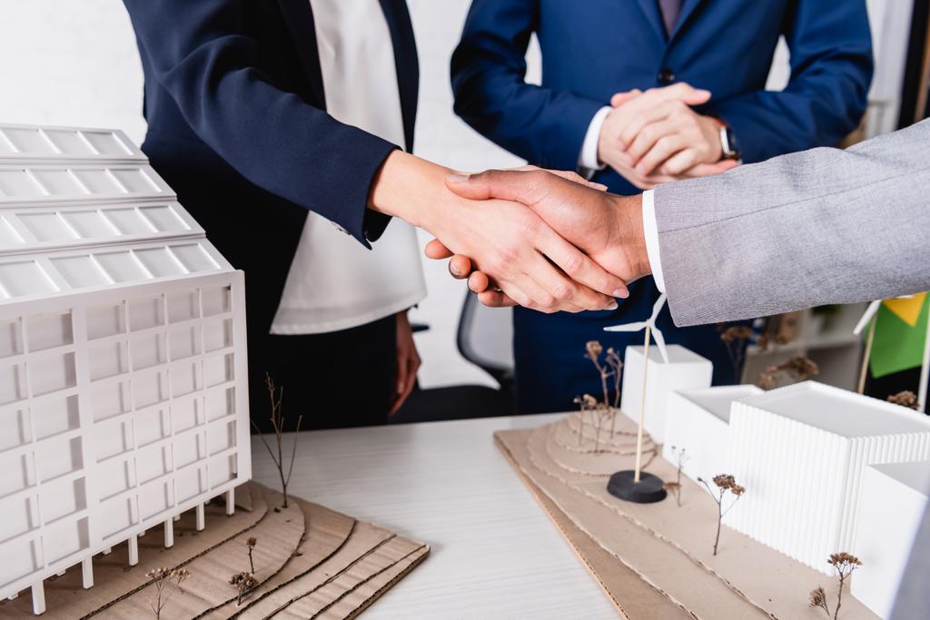 częściowy widok międzyrasowych partnerów biznesowych ściskających dłonie w pobliżu modelu budynku z alternatywną elektrownią - Zdjęcie, obraz